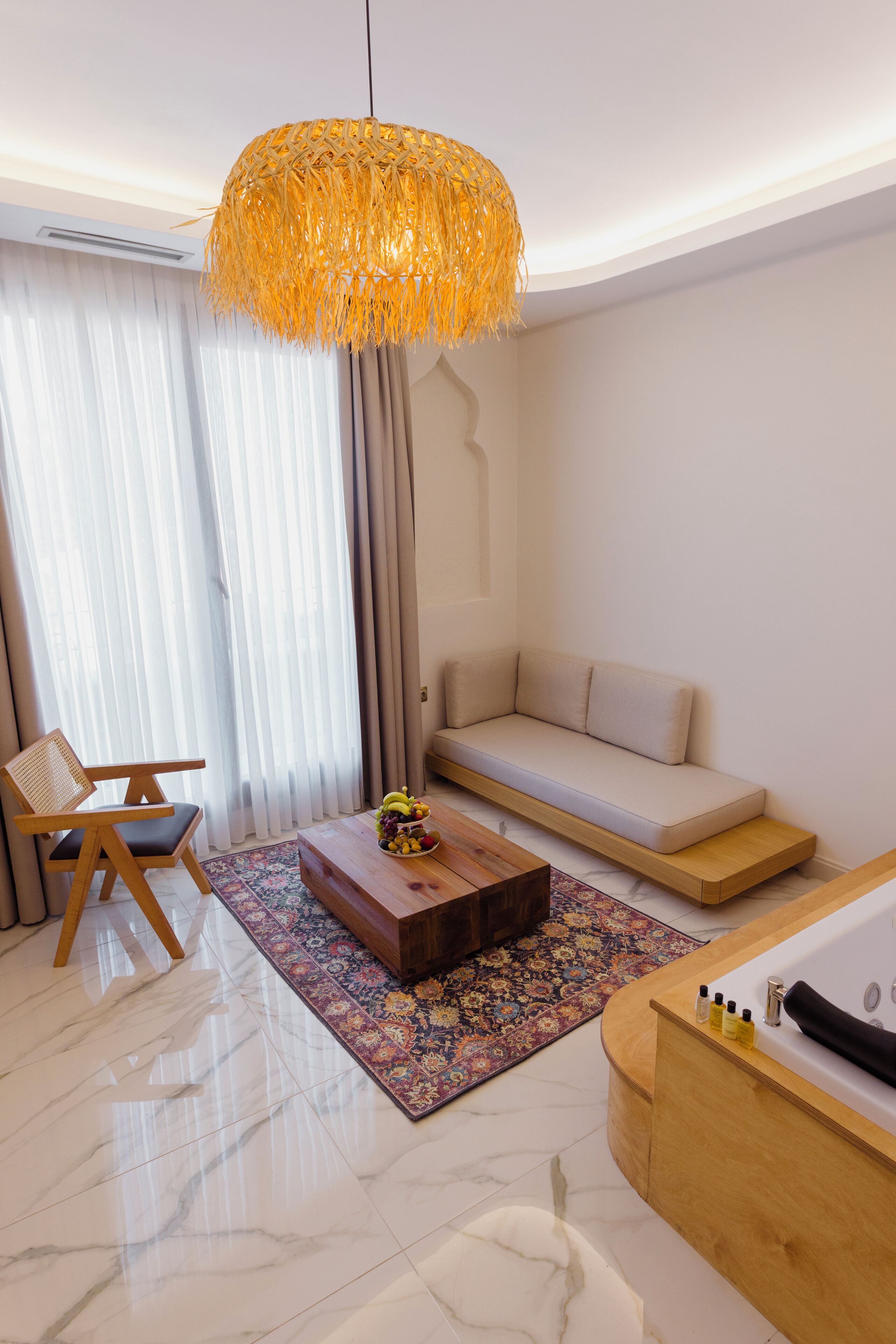 A modern hotel room with a wooden sofa, a wooden coffee table with fruit, a large hanging light fixture, a bathtub, and a patterned rug.