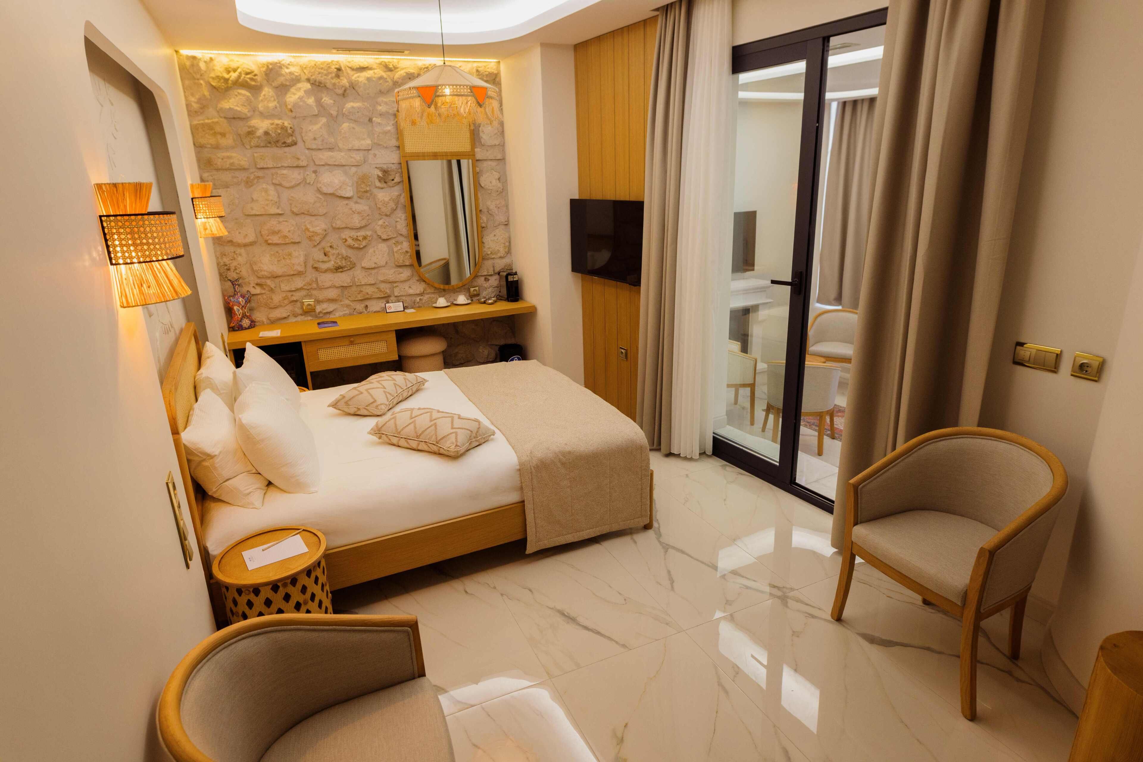 Una habitación de hotel moderna con una pared de acento de piedra, una cama con ropa de cama blanca, una mesita de noche de madera, un televisor y un balcón con cortinas.