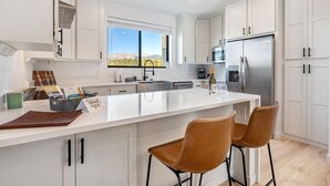 A modern kitchen with white cabinetry, a large island with bar stools, a stainless steel refrigerator, and a window with a view. - Fraser Ridge Retreat | 3BR/3.5BA, Free WP Shuttle (Fraser)