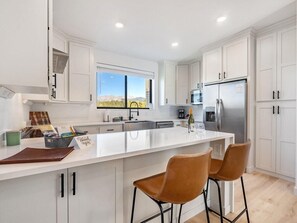 A modern kitchen with white cabinetry, a large island with bar stools, a stainless steel refrigerator, and a window with a view. - Fraser Ridge Retreat 3 Bed 3.5 Bath - Sleeps 9 (Fraser)