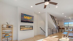 A modern living room with a fireplace, a mounted TV, a ceiling fan, and a staircase leading to a kitchen area. - Fraser Ridge Retreat | 3BR/3.5BA, Free WP Shuttle (Fraser)