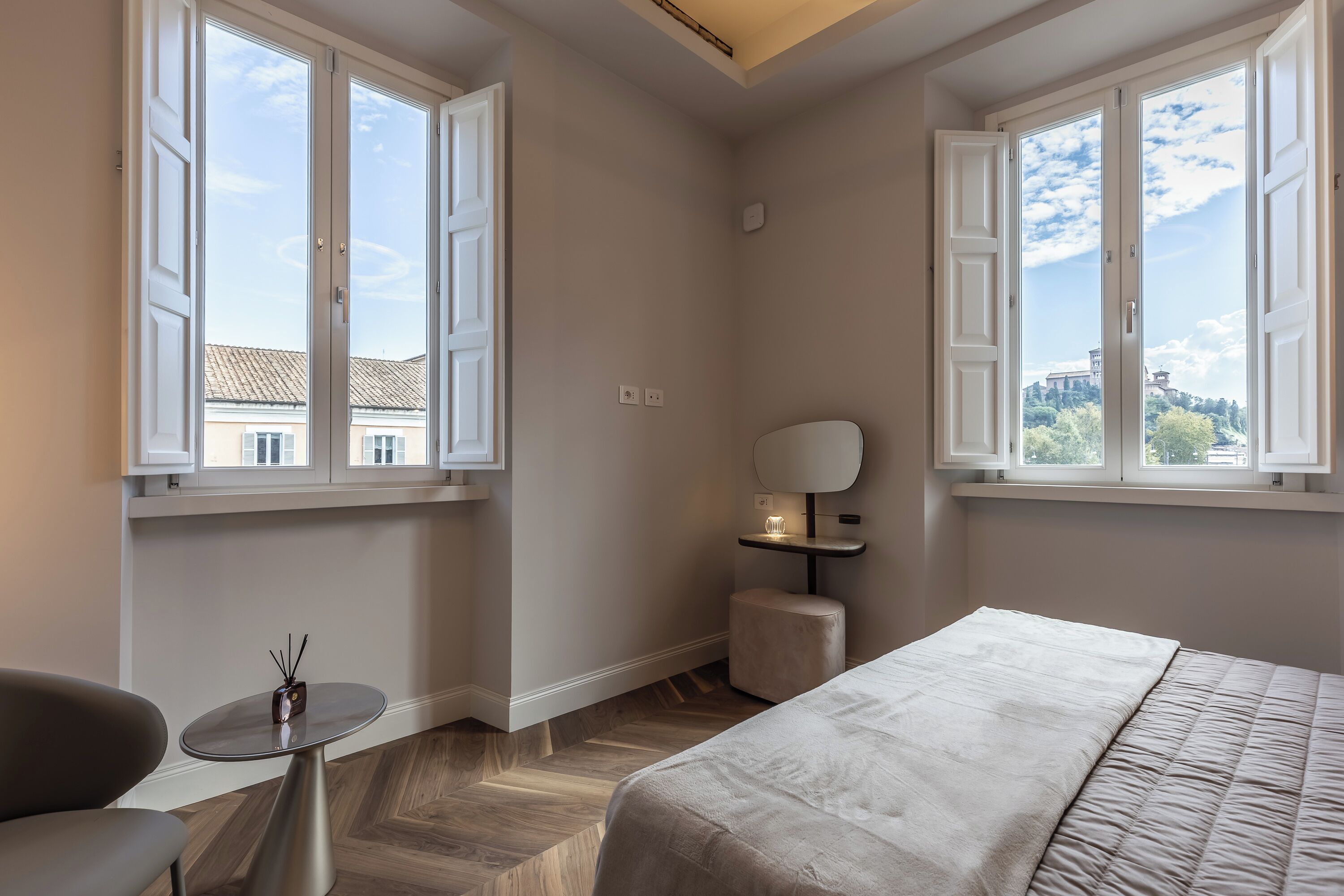 A bedroom with a bed, a chair, a small table, and a nightstand. There are two windows providing a view of buildings and trees.