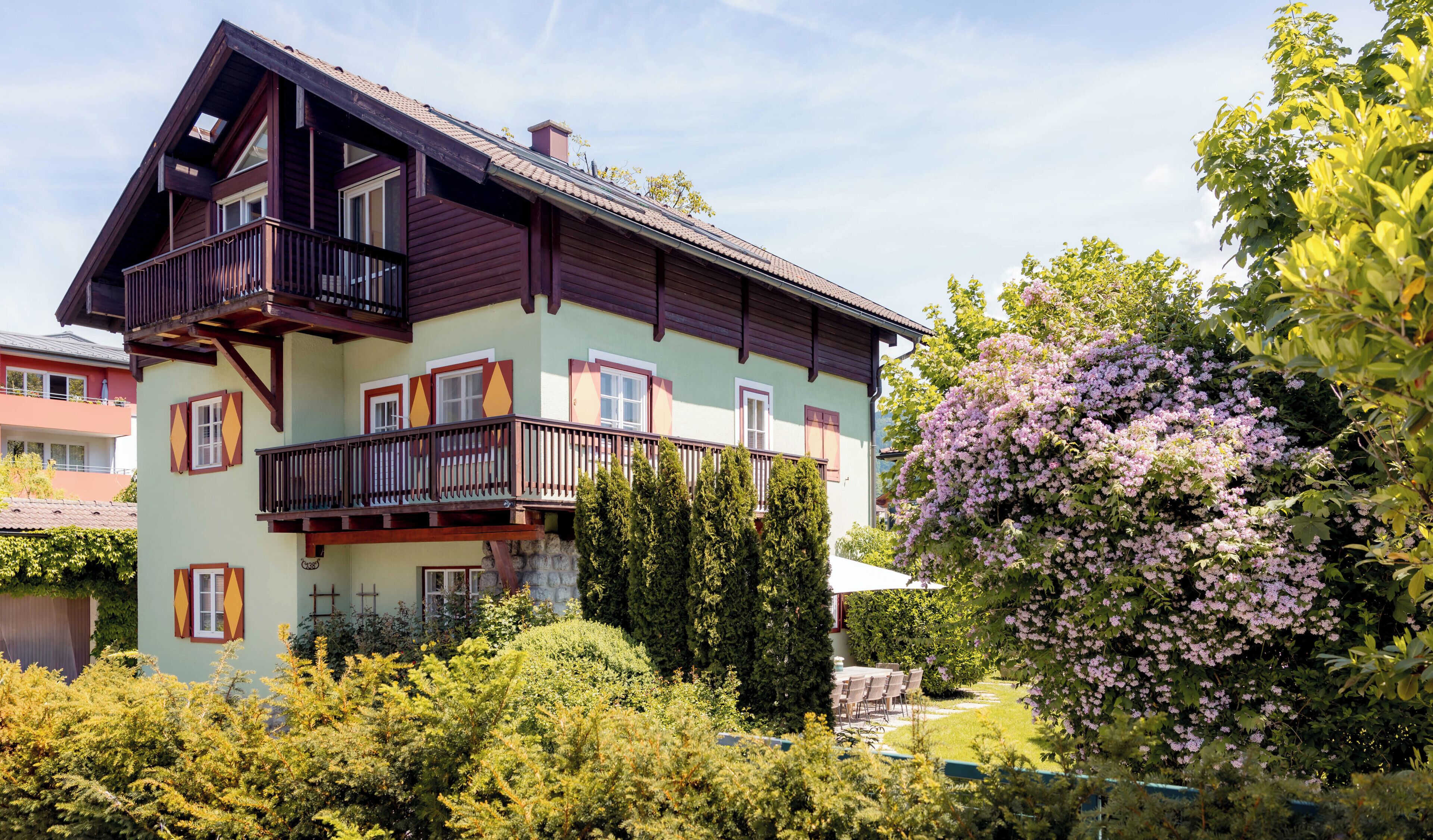 Une maison à plusieurs étages dotée d’un balcon, entourée d’une végétation luxuriante et d’arbres en fleurs.