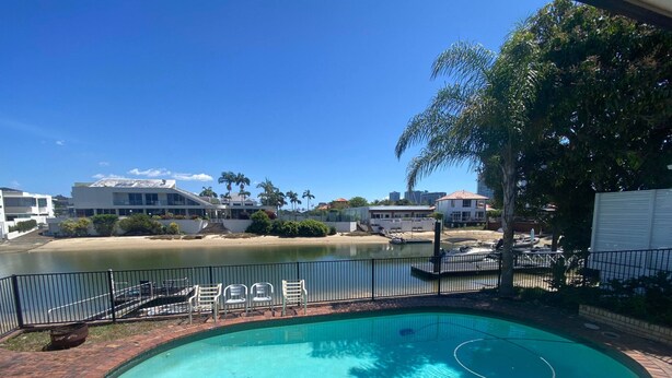 A swimming pool with a brick border, surrounded by a fence, and overlooking a beach with palm trees and buildings in the background.