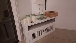 A white console table with a lamp, candles, a remote control, and a wooden basket.
