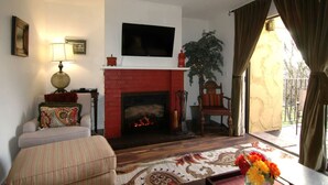 Una sala de estar con chimenea, televisor montado en la pared, sofá, sillón y una mesa con flores.