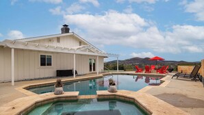 Une piscine avec une maison en arrière-plan, un espace patio avec des chaises rouges et une vue sur les montagnes.