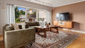 Een woonkamer met een bank, salontafel en televisie. Er is een groot raam met gordijnen, een kamerkleed met een patroon en een houten kast.