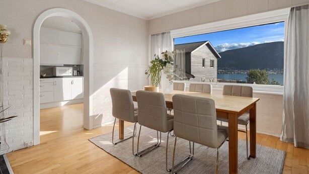Een moderne eetruimte met een grote tafel en stoelen, uitzicht op een berg en meer door een groot raam, en een keuken op de achtergrond.
