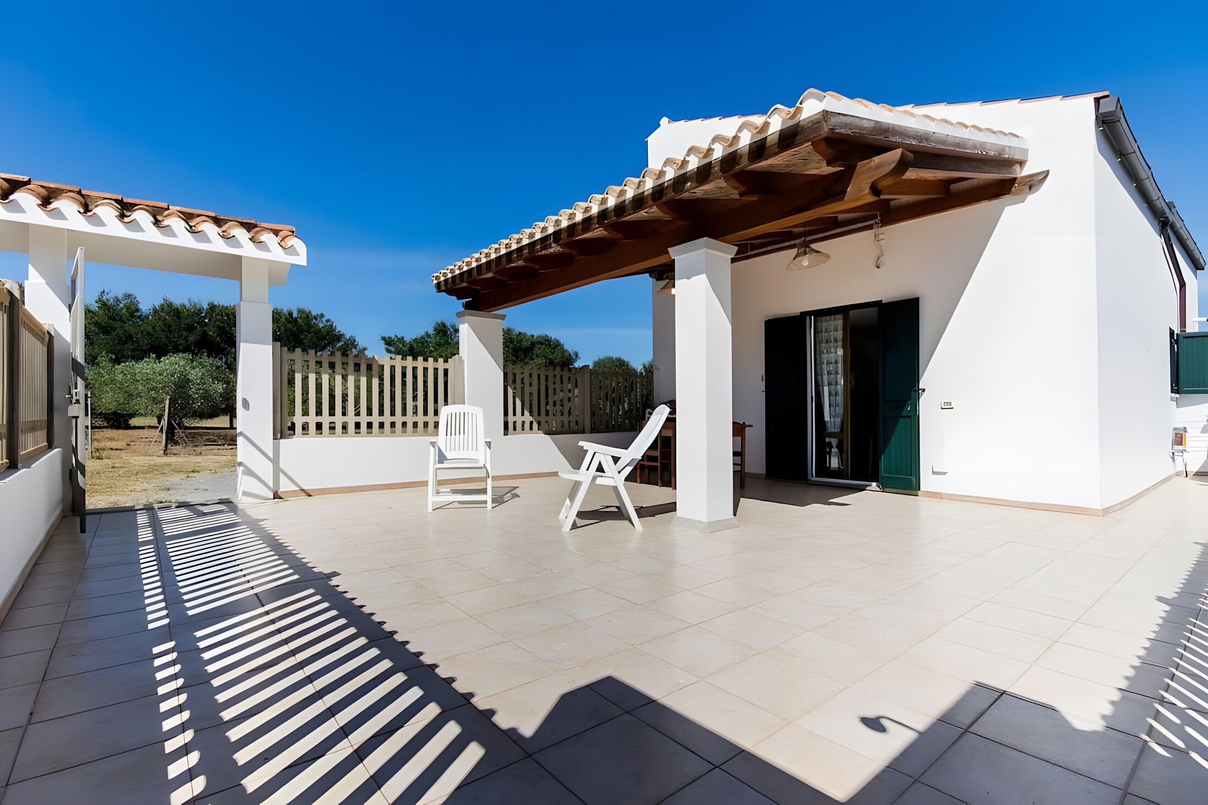 Eine überdachte Terrasse mit zwei weißen Stühlen, einer Pergola und einem Gebäude mit weißen Fliesen und einem Ziegeldach.