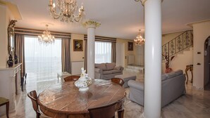 Uma sala de estar espaçosa com uma mesa de jantar redonda de madeira, um sofá cinzento e uma escada para um andar superior.