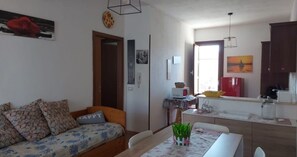 Un salon avec un canapé, une table à manger avec des chaises, un coin cuisine avec un réfrigérateur et une cuisinière, et une porte donnant sur une autre pièce.