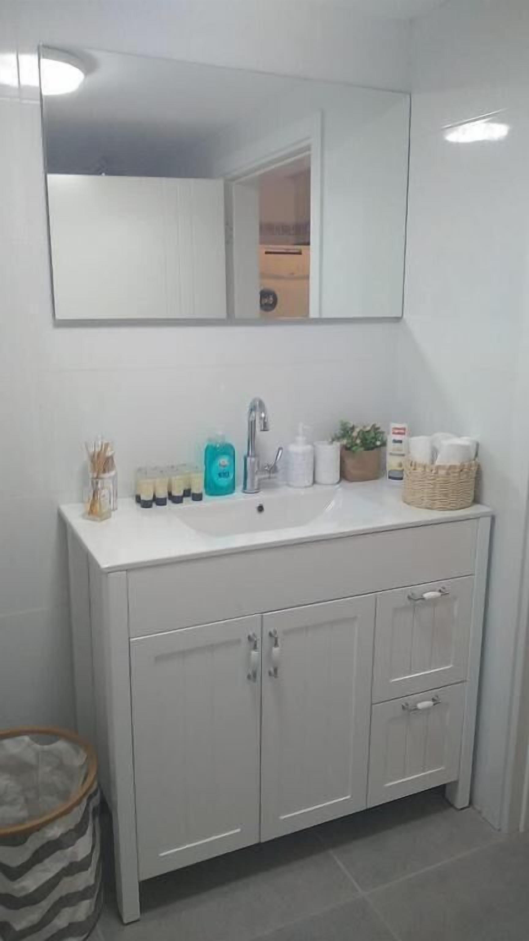 A bathroom with a white sink, a mirror above it, and a cabinet below. There are toiletries on the sink and a basket on the cabinet.