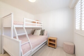A bunk bed with a ladder, a small wooden nightstand, and a round ottoman in a room with white walls and a window with blinds. - Seaspray Sands Coastal Retreat in Nelson Bay (Nelson Bay)
