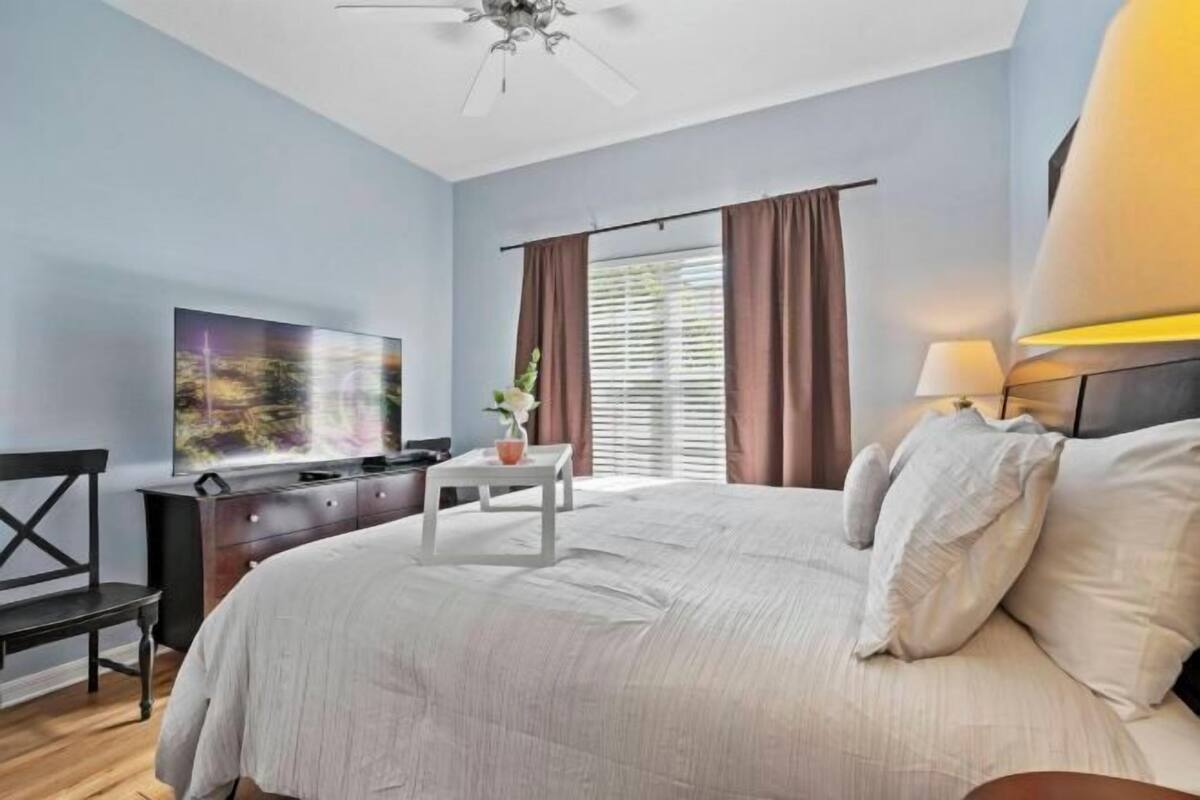 Un dormitorio con cama, televisión, escritorio, silla y ventana con cortinas.
