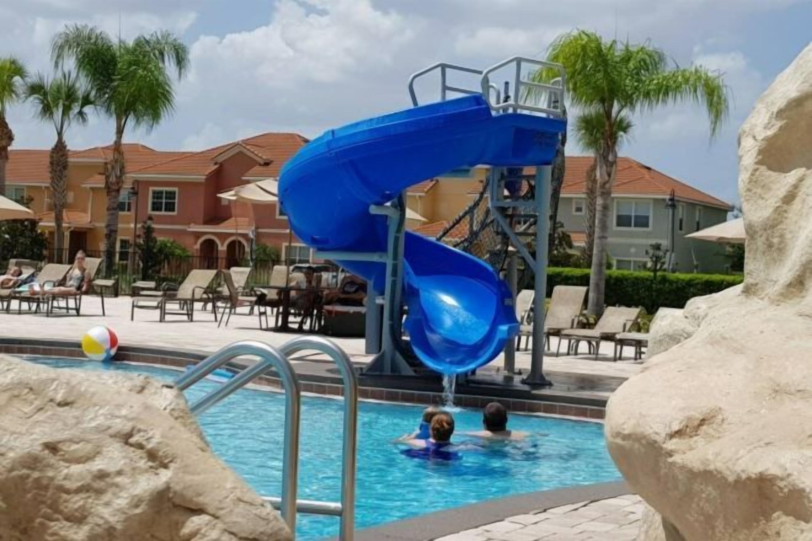 A swimming pool with a blue water slide, lounge chairs, palm trees, and a building in the background.