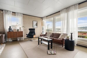 Ein modernes Wohnzimmer mit einer Couch, einem schwarzen Sessel, einem Holzschrank und einem großen Fenster mit Ausblick.