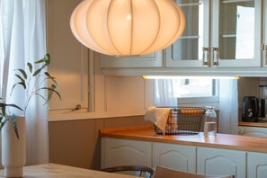 Eine Küche mit weißen Schränken, einer Holzausziehtafel, einer Pendelleuchte und einem Fenster mit Vorhängen.