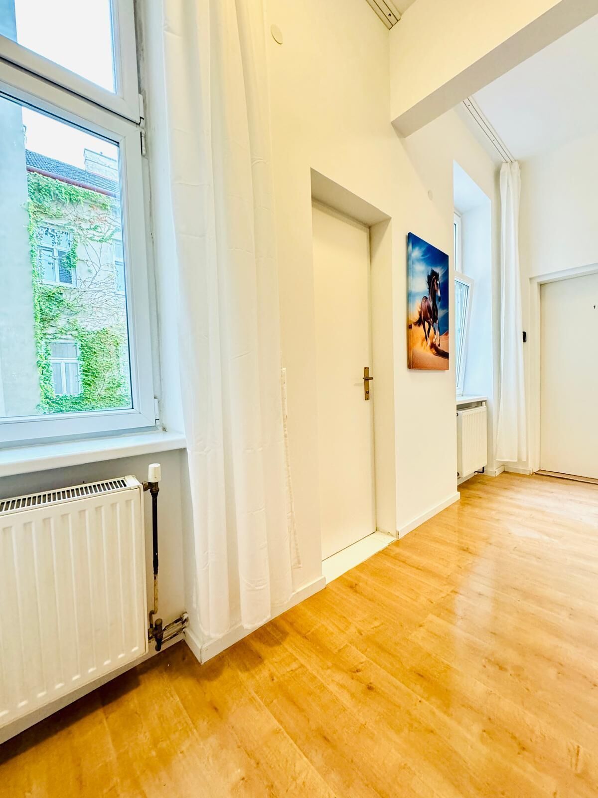 En rummelig gang med trægulv, hvide vægge og en radiator. Der er et vindue til venstre og en dør i enden af gangen.