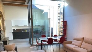 Ein modernes Wohnzimmer mit Sofa, Hocker und einem roten Teppich. Es gibt einen Essbereich mit Tisch und Stühlen sowie eine Küche im Hintergrund.