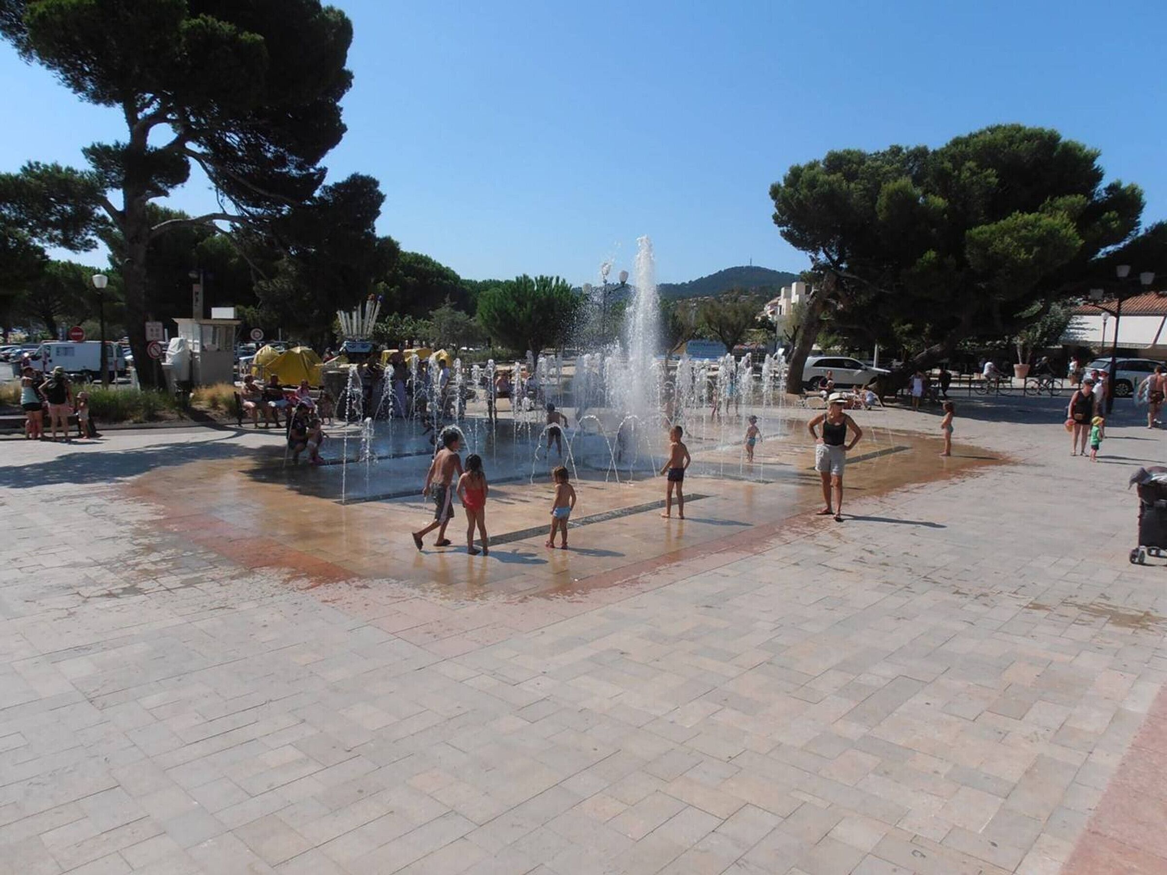 Uma praça pública com uma fonte central, rodeada por árvores e pessoas.
