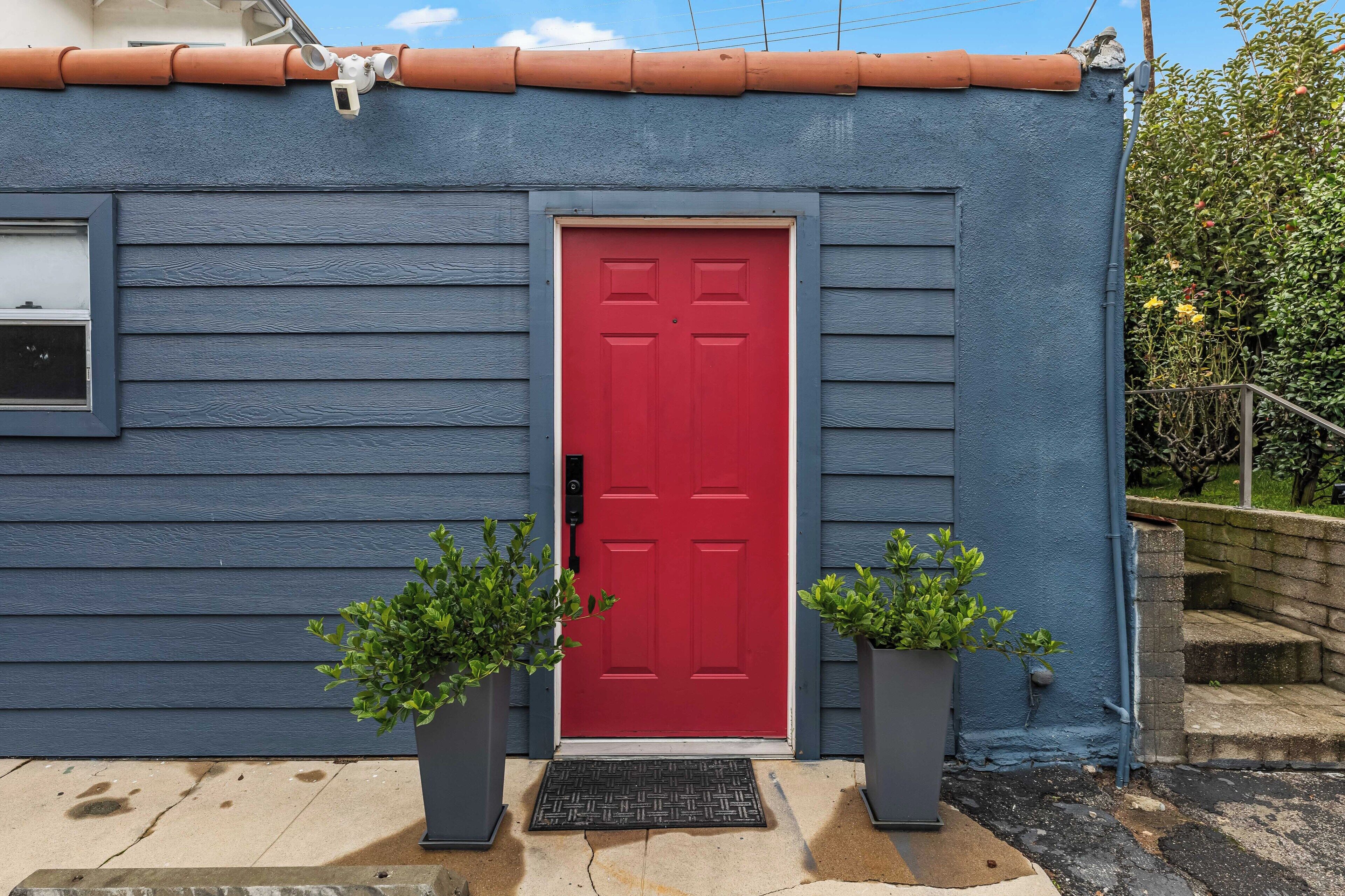 A blue house with a red door, two potted plants, and a metal doormat.