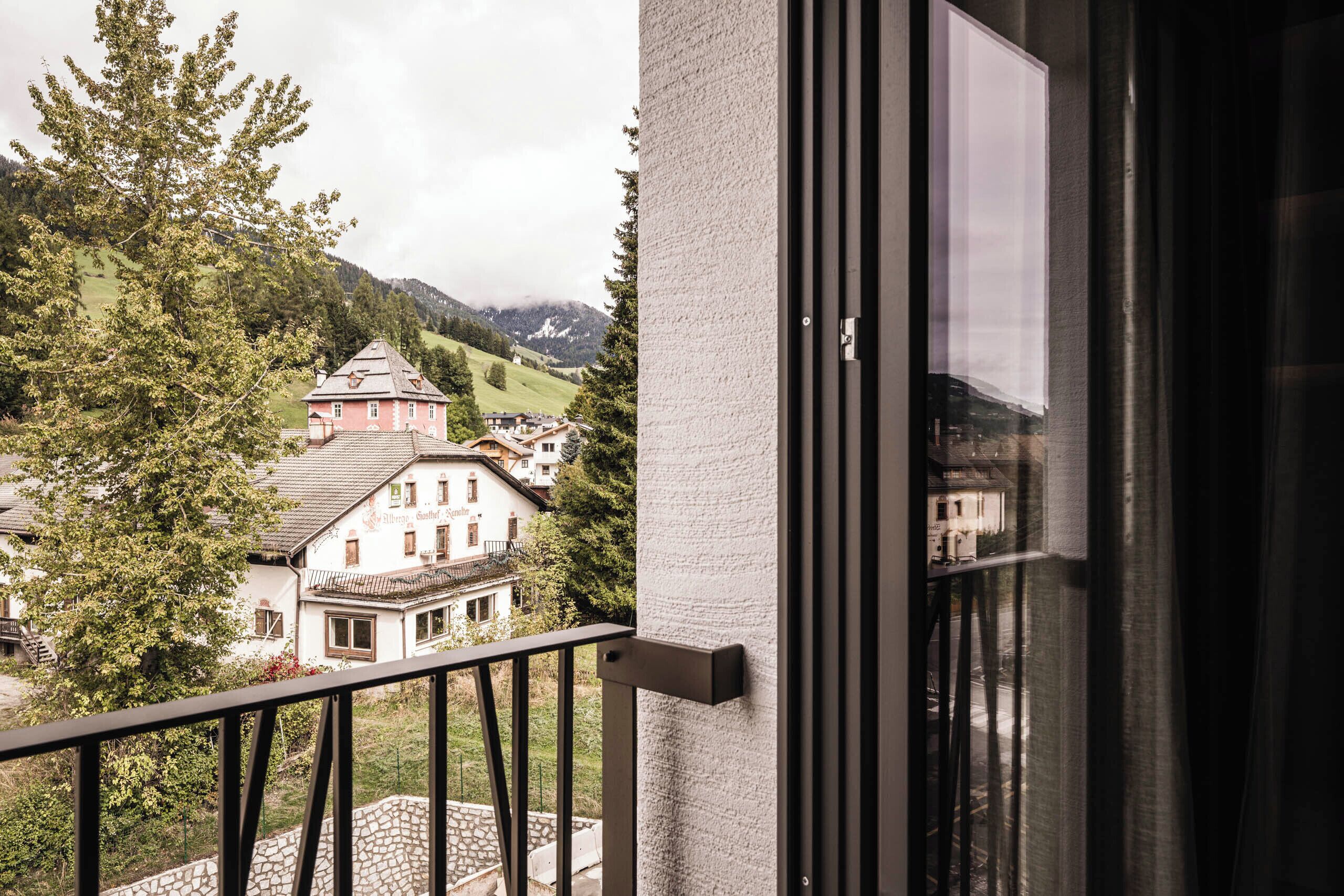 Un balcon doté d’un mur en pierre et d’une balustrade en métal, offrant une vue sur un village aux bâtiments traditionnels et aux collines verdoyantes.
