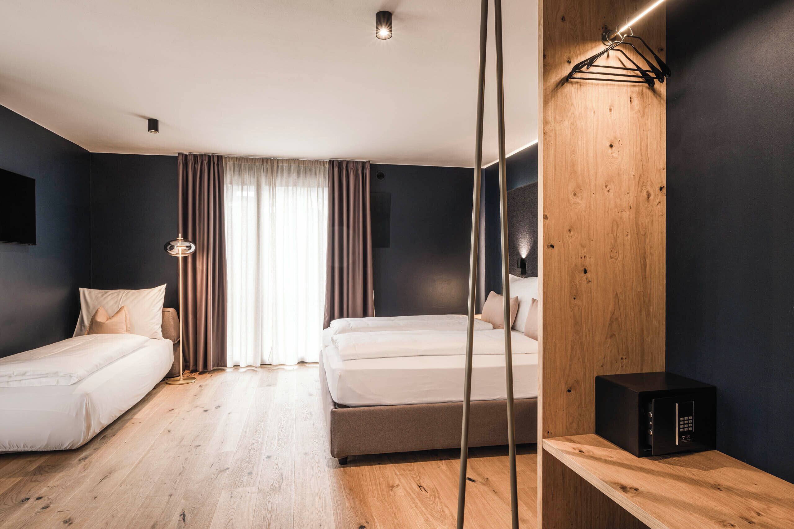 A modern hotel room with a wooden floor, a large bed, a smaller bed, a bedside table, a mirror, and a wall-mounted TV.
