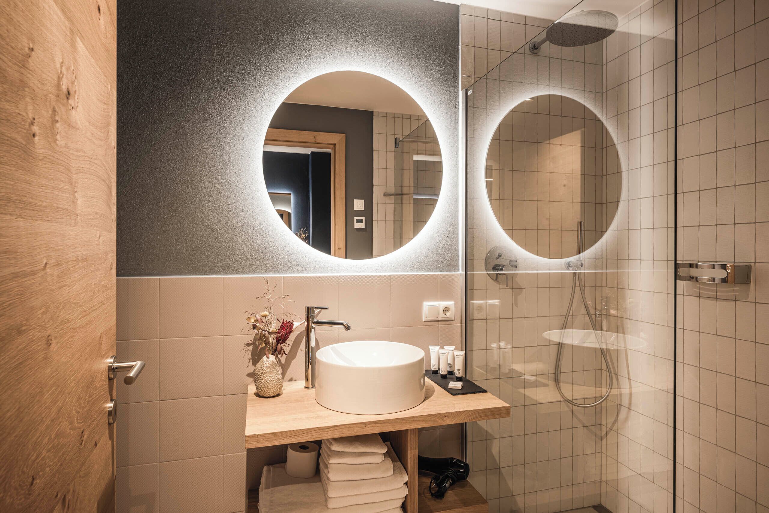 Une salle de bain moderne avec un meuble-lavabo en bois, un miroir rond et une douche à parois de verre.