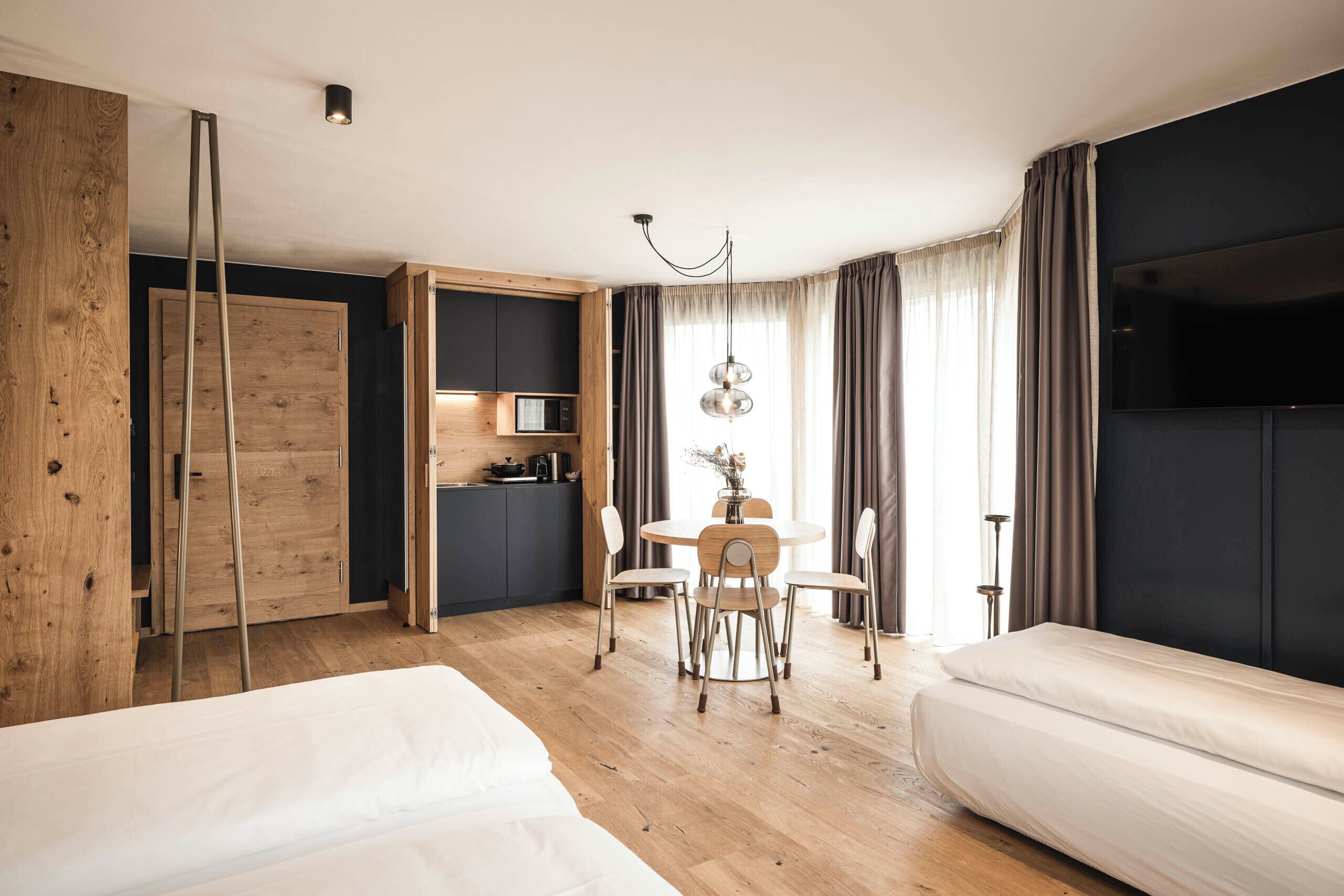 A modern hotel room with a wooden floor, a bed, a dining table with chairs, a kitchen area, and a TV.