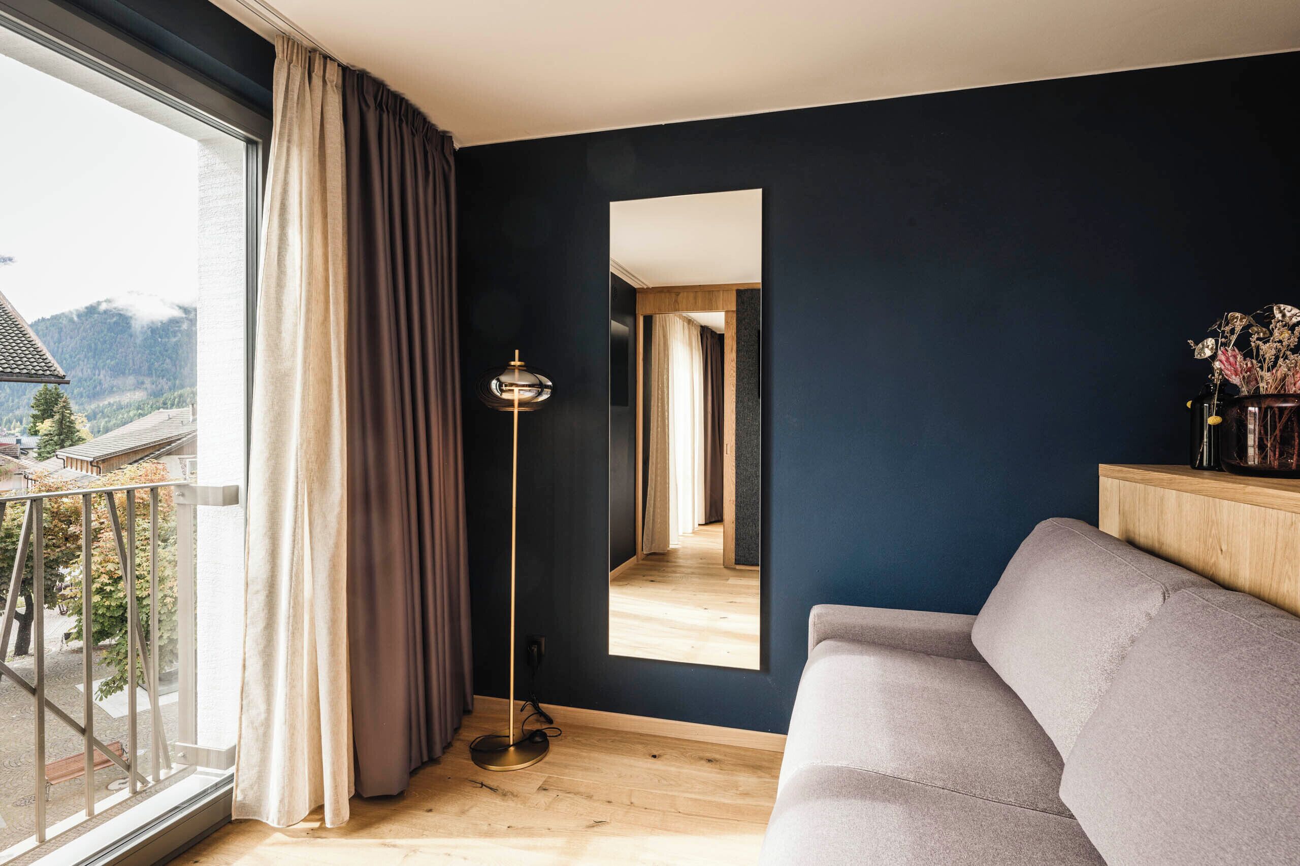 Een moderne woonkamer met een groot raam, een bank, een vloerlamp en een spiegel die de kamer reflecteert.