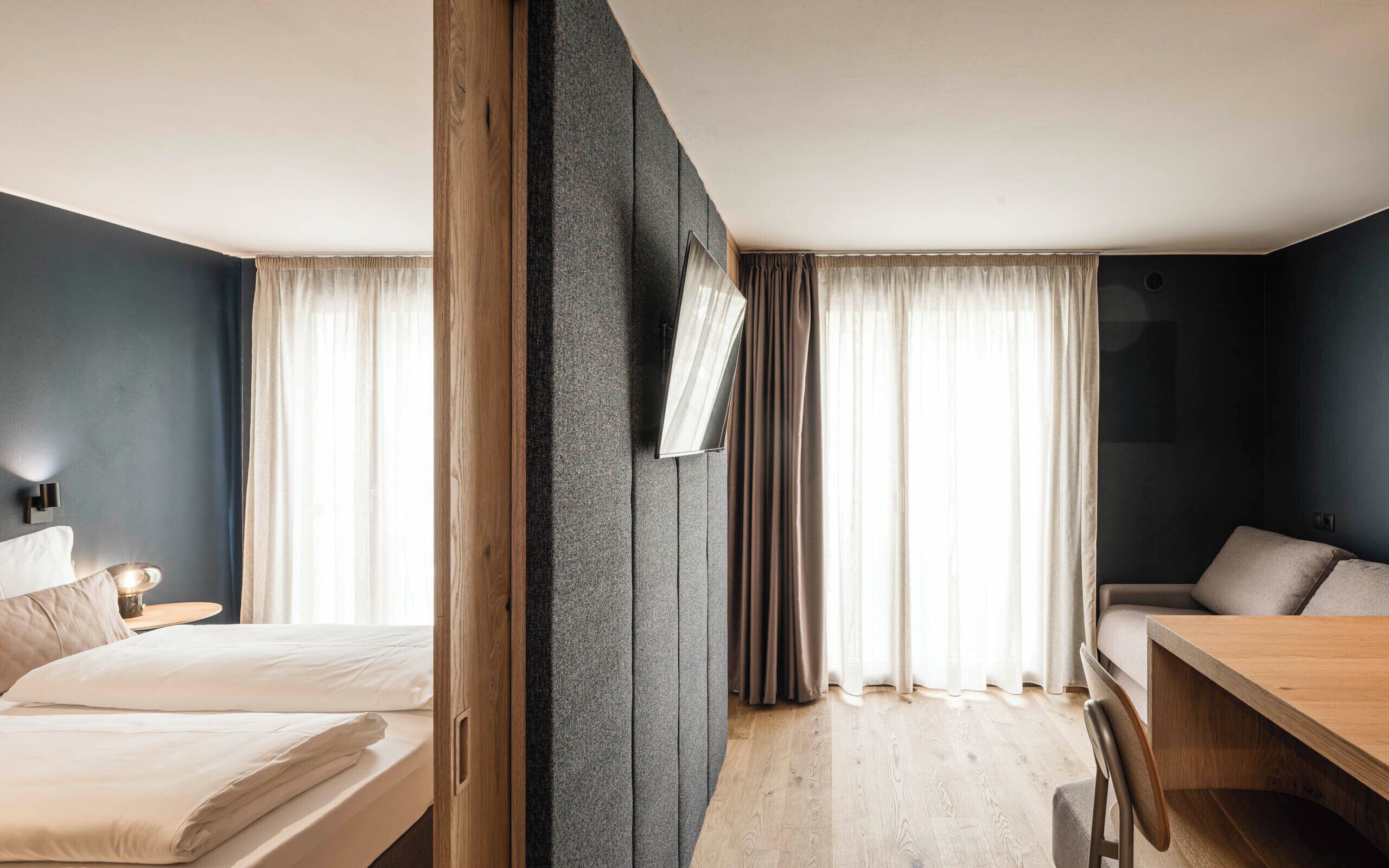 A modern hotel room with a bed, a desk, and a chair. There are curtains on the windows, and a mirror is mounted on the wall.