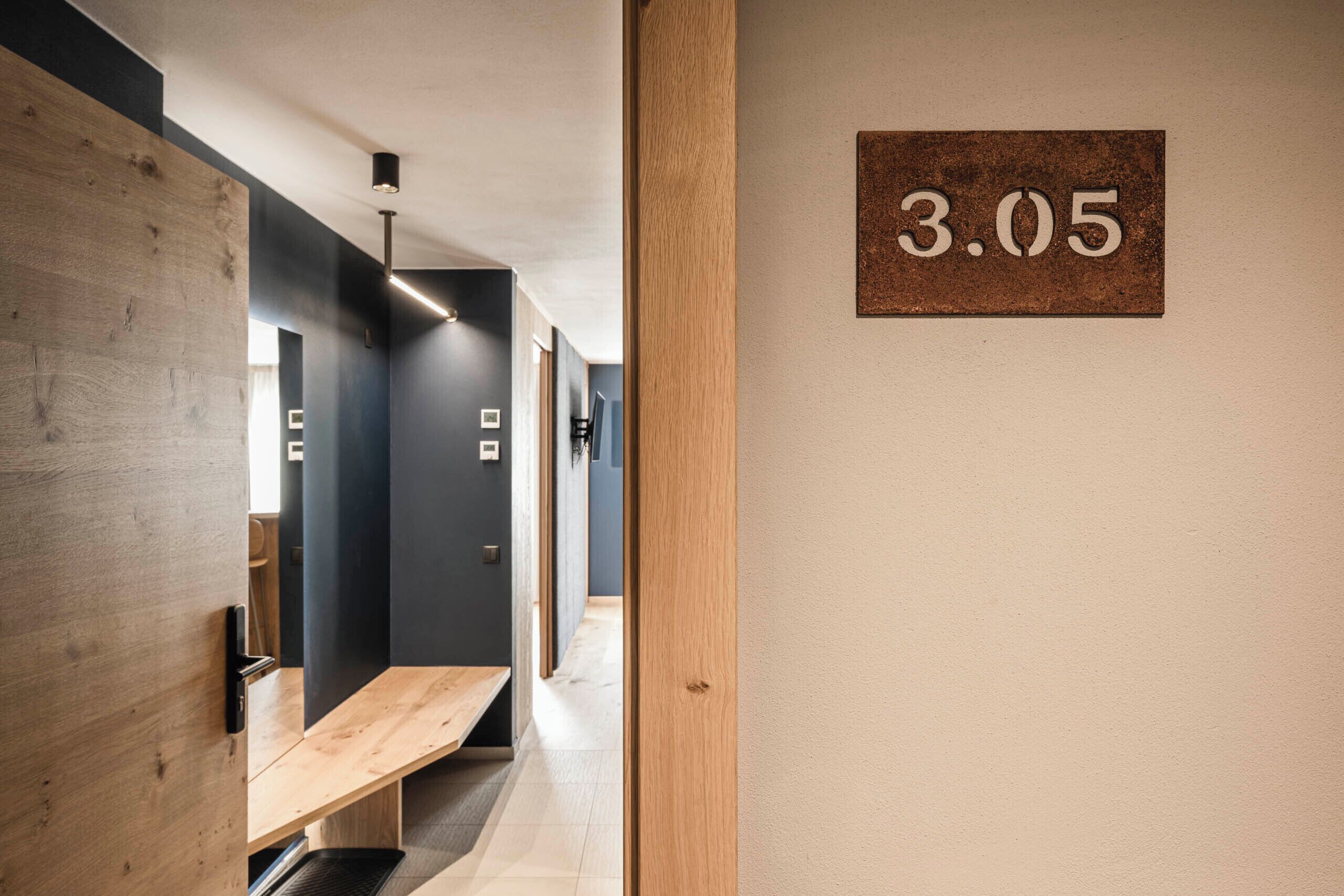 A hallway with a wooden bench, a number sign '3.05' on the wall, and a modern light fixture.