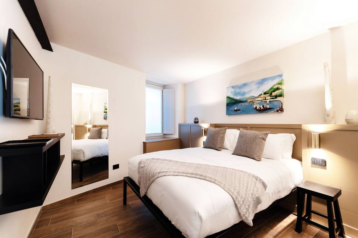 Une chambre à coucher moderne avec un grand lit, une table de chevet, un miroir et un tableau représentant un paysage côtier.