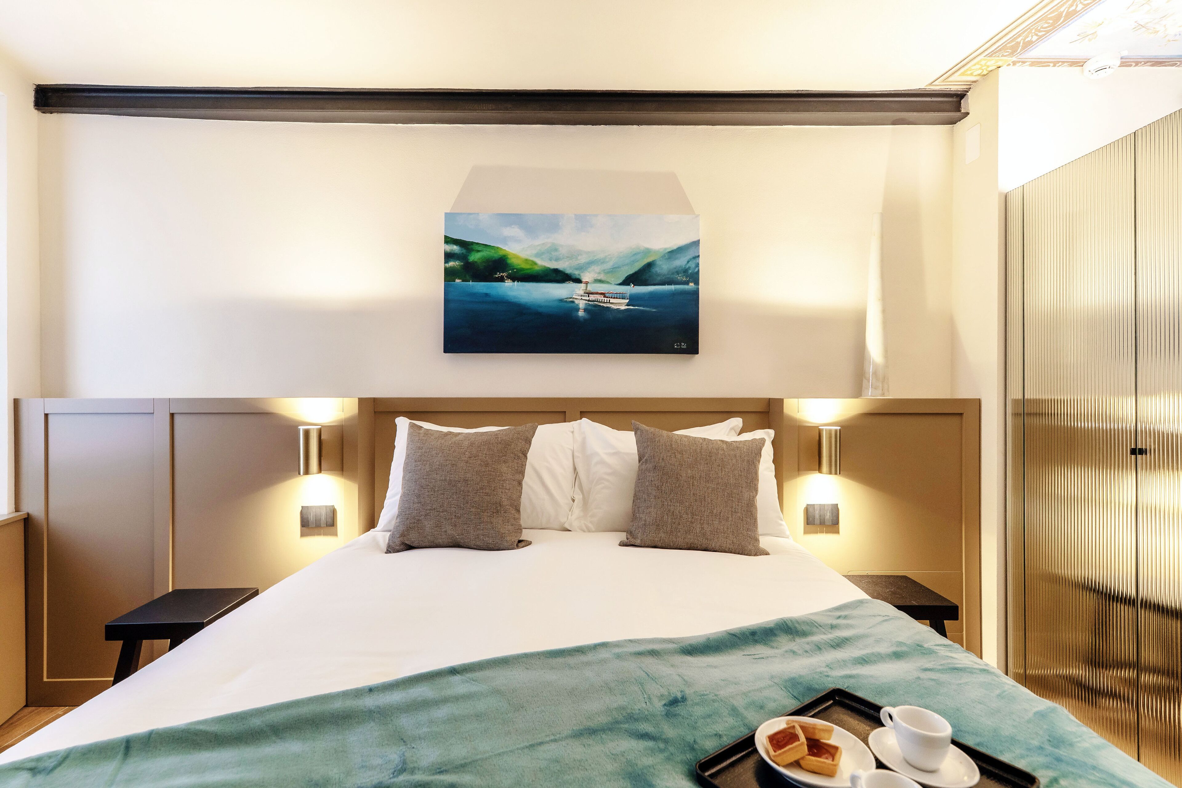 A hotel room with a bed, pillows, a painting of a lake, and a tray with breakfast items.