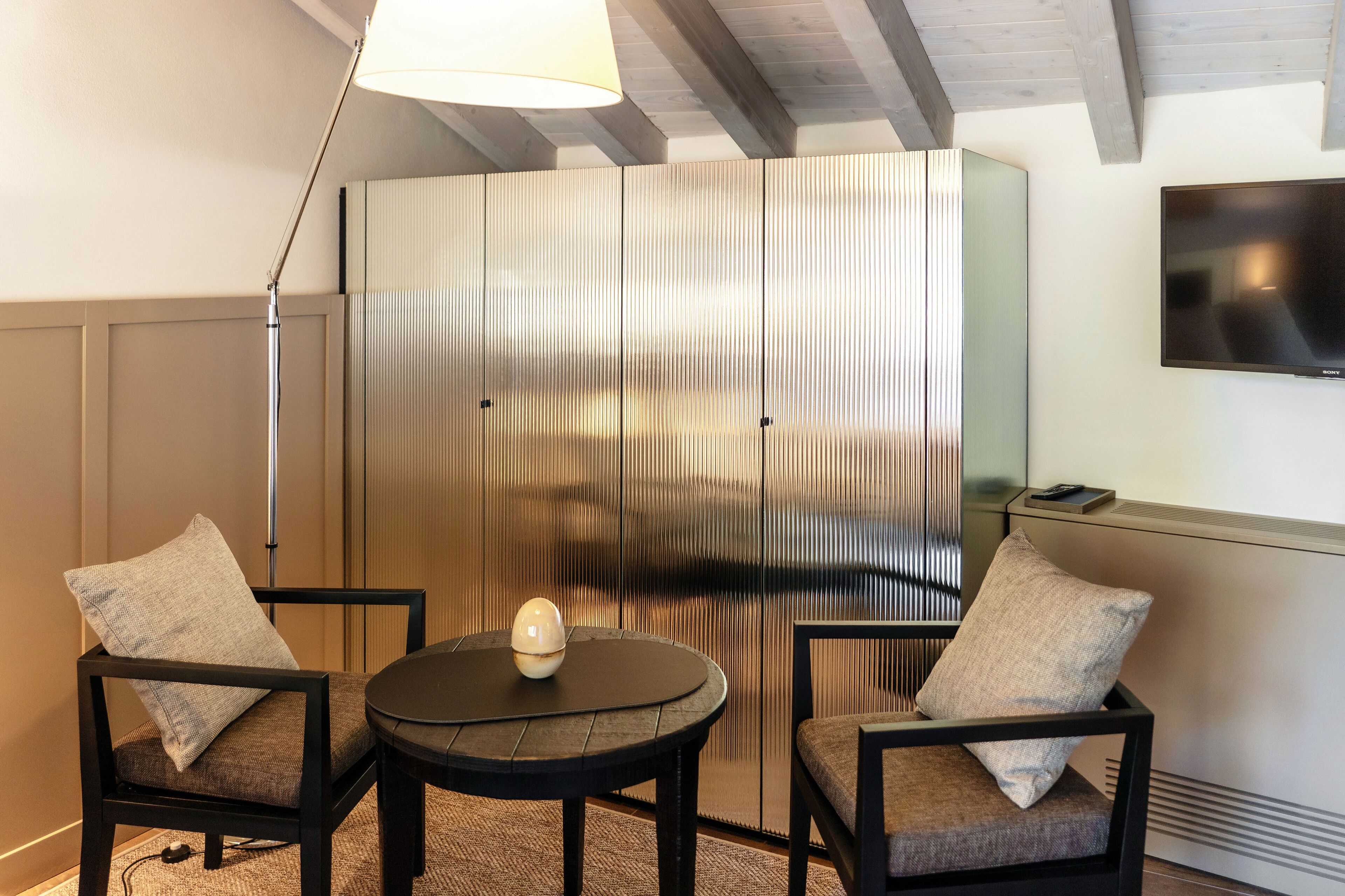 Una habitación moderna con una mesa redonda, dos sillas, una lámpara y un panel metálico grande.
