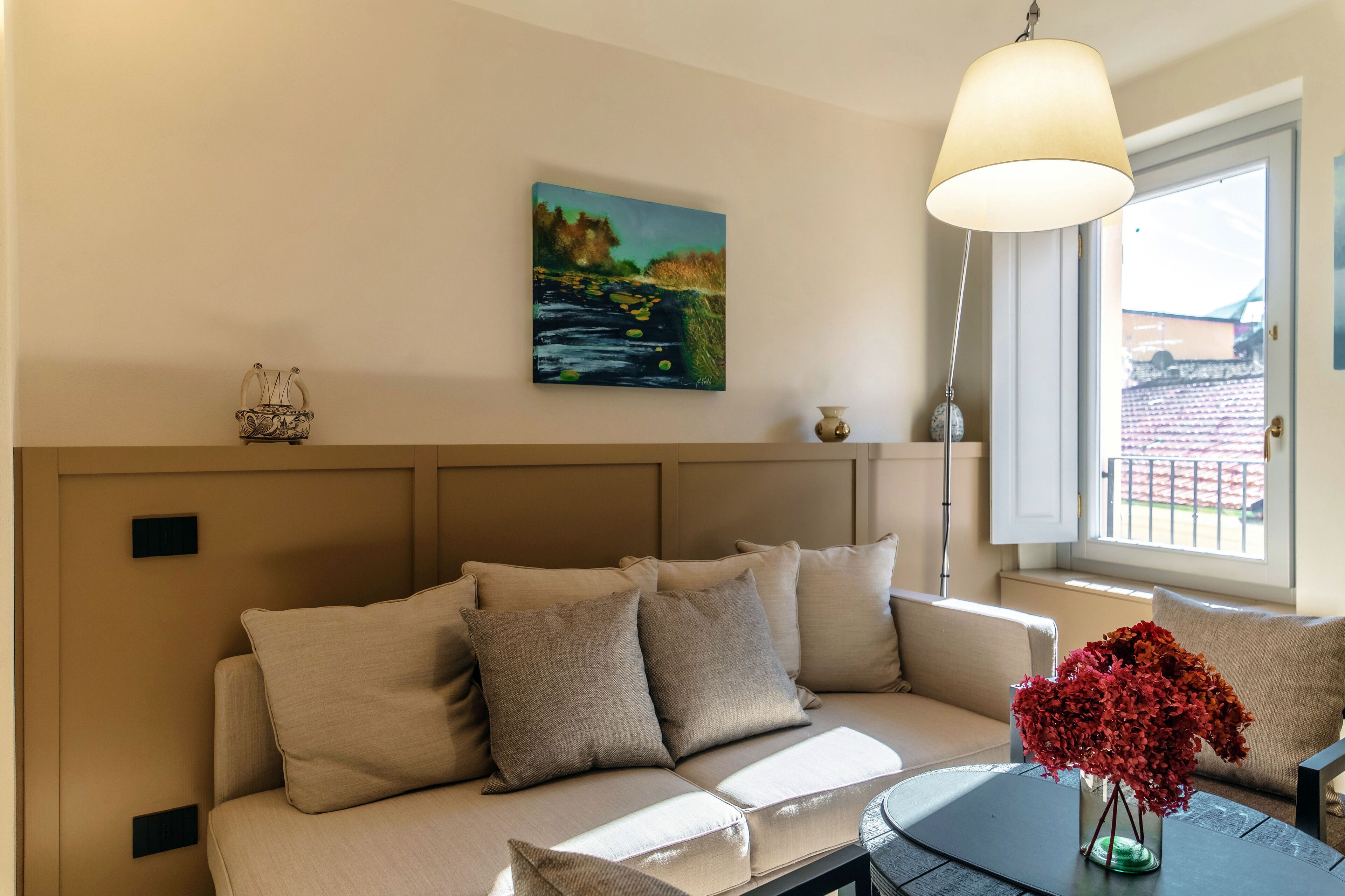 A modern living room with a beige sofa, a painting on the wall, and a vase with red flowers on a side table.