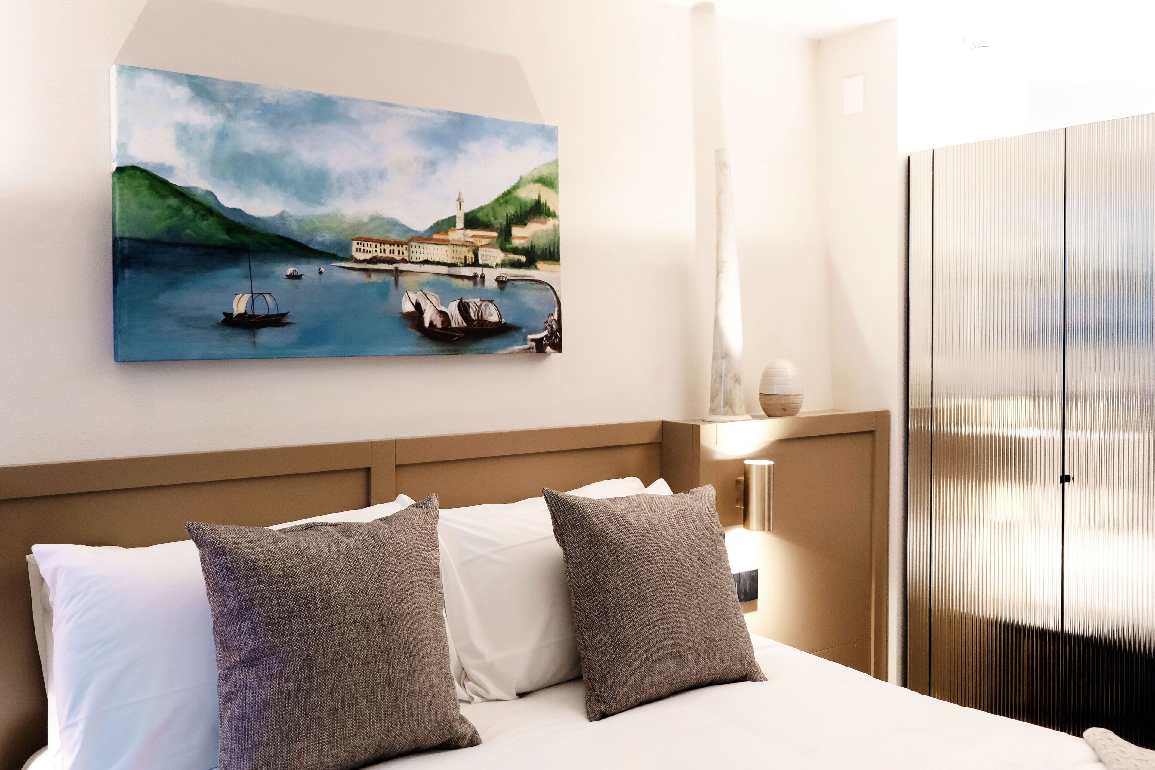 Quarto de hotel com cama, almofadas, um quadro de uma paisagem costeira e um guarda-roupa.