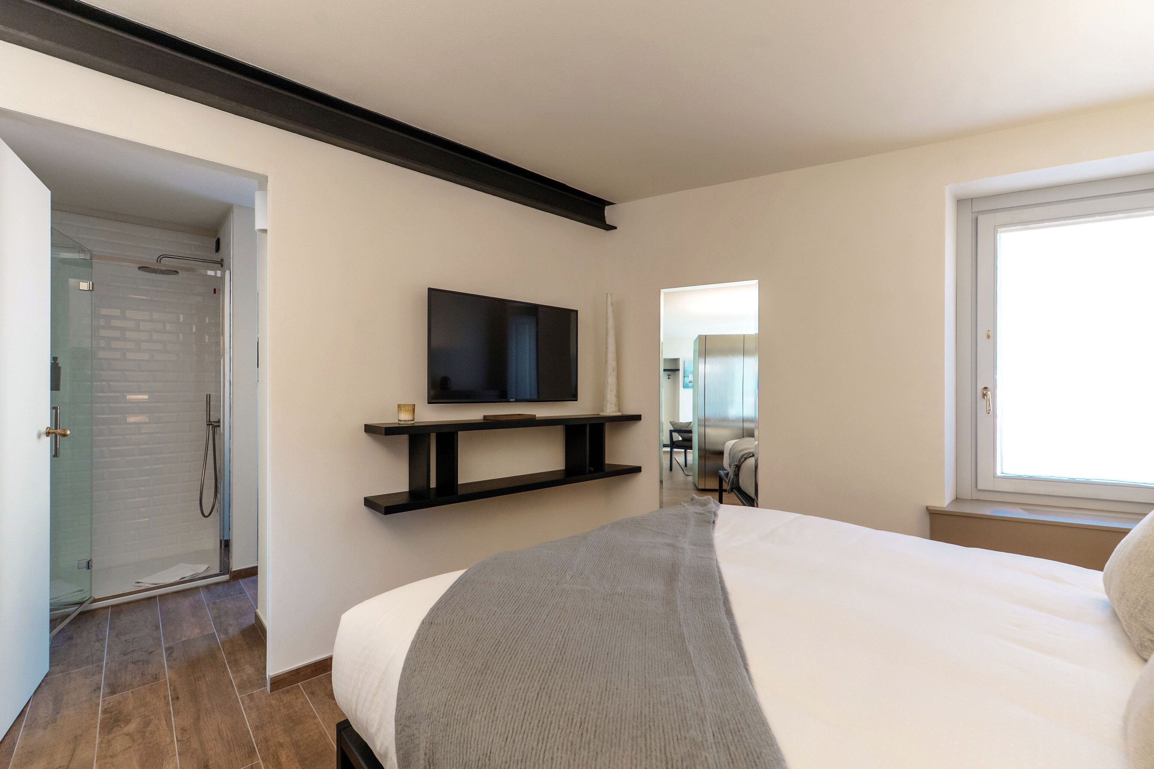 A modern hotel room with a large bed, a TV mounted on the wall, a shower area, and a window providing natural light.