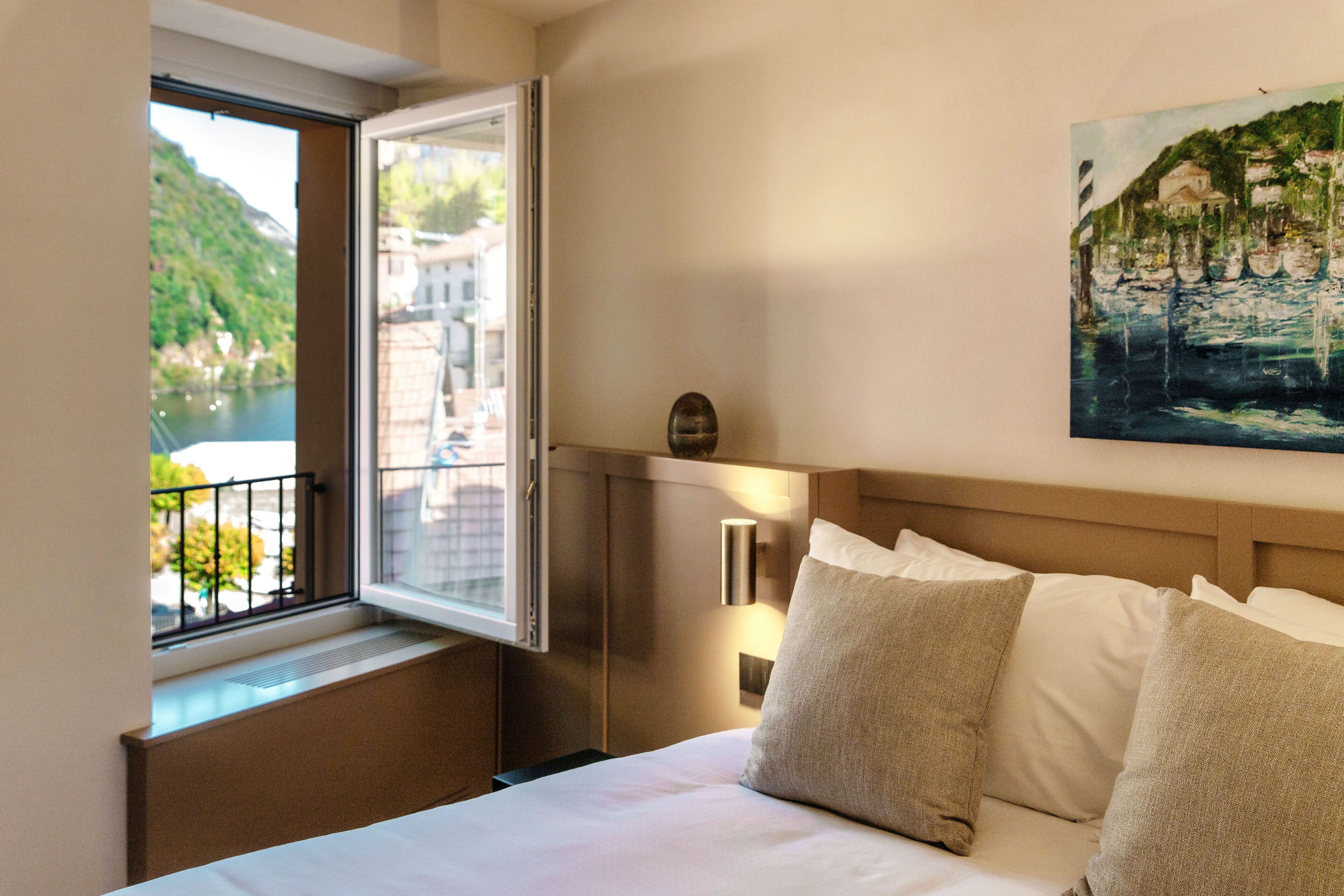 A hotel room with a bed, a window with a view of a river and buildings, and a painting on the wall.