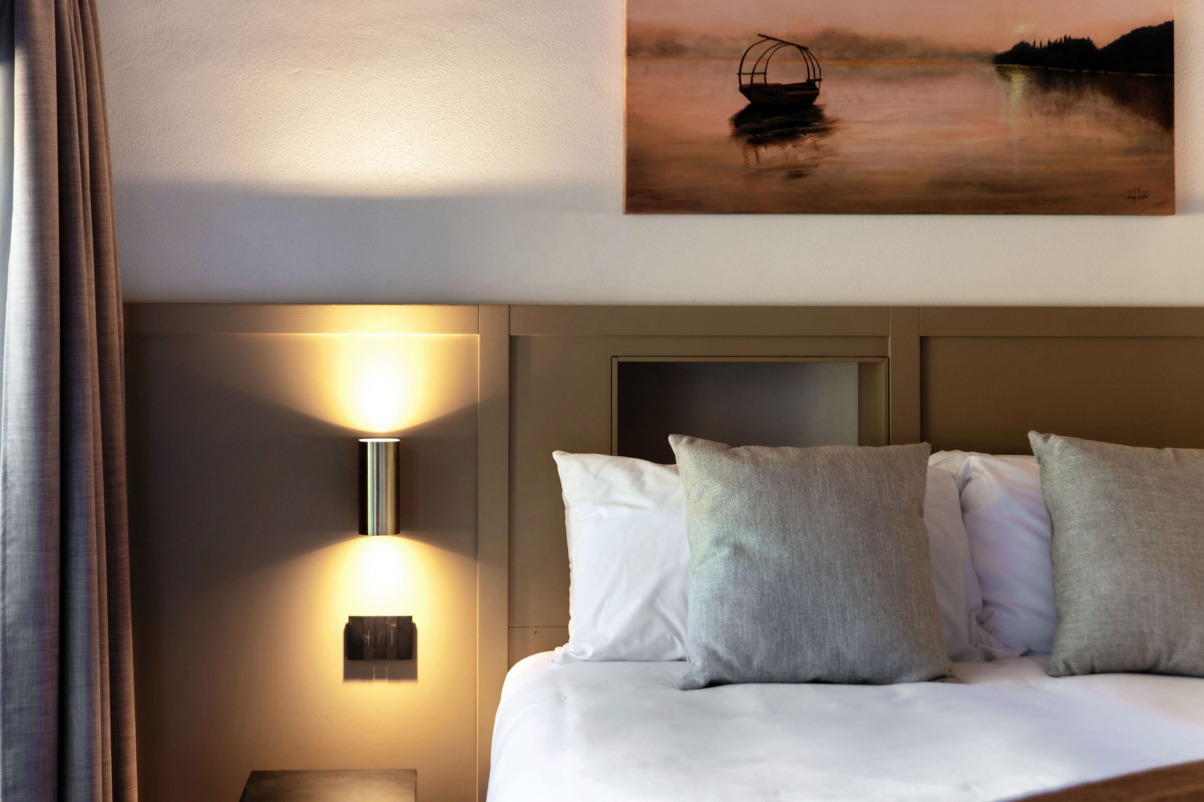 A neatly made bed with white and grey pillows, a wall-mounted light fixture, and a painting of a boat on water.