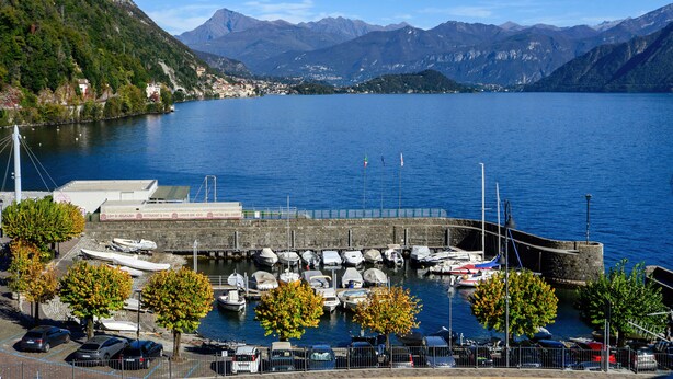 A marina with boats docked, a parking area with cars, and a backdrop of mountains. - Home Holidays (Argegno)