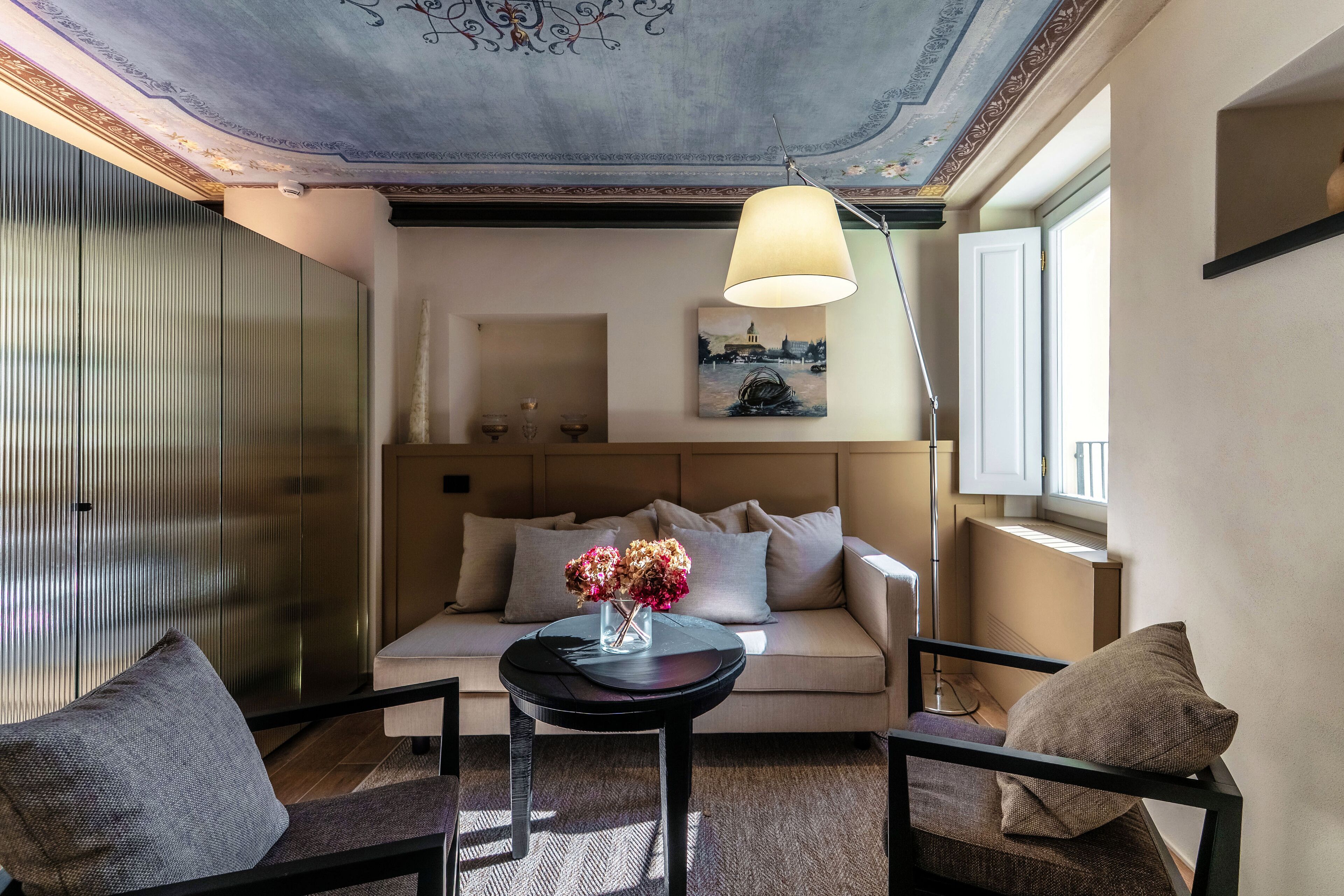 Een moderne woonkamer met een bank, twee fauteuils, een salontafel met een bloevas, een schilderij aan de muur en een plafond met ingewikkelde decoraties.