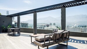 Una terraza en la azotea con un área de estar exterior moderna, una pérgola y vistas a un paisaje urbano con montañas al fondo.