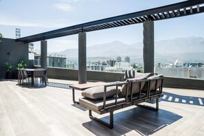 Eine Dachterrasse mit einer modernen Sitzgelegenheit im Freien, einer Pergola und Blick auf eine Stadtlandschaft mit Bergen im Hintergrund.