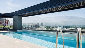 Una piscina en la azotea con agua azul cristalina, barandillas metálicas y vistas a un paisaje urbano y montañas al fondo.