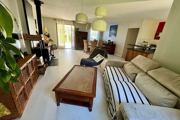 A spacious living room with a sofa, a wooden coffee table, and a dining area with chairs. - Spacious villa near the beach, in a quiet wooded setting (Lanton)