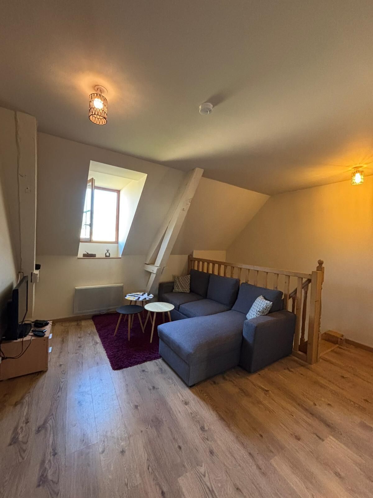 Une pièce de style « loft » avec un plafond en pente, un escalier en bois intégré, un canapé d’angle gris, une petite table ronde et une fenêtre laissant entrer la lumière naturelle.