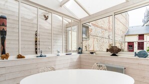 Una habitación con una mesa redonda grande, sillas blancas, un ventanal con vista a un edificio y varios objetos decorativos en el alféizar.