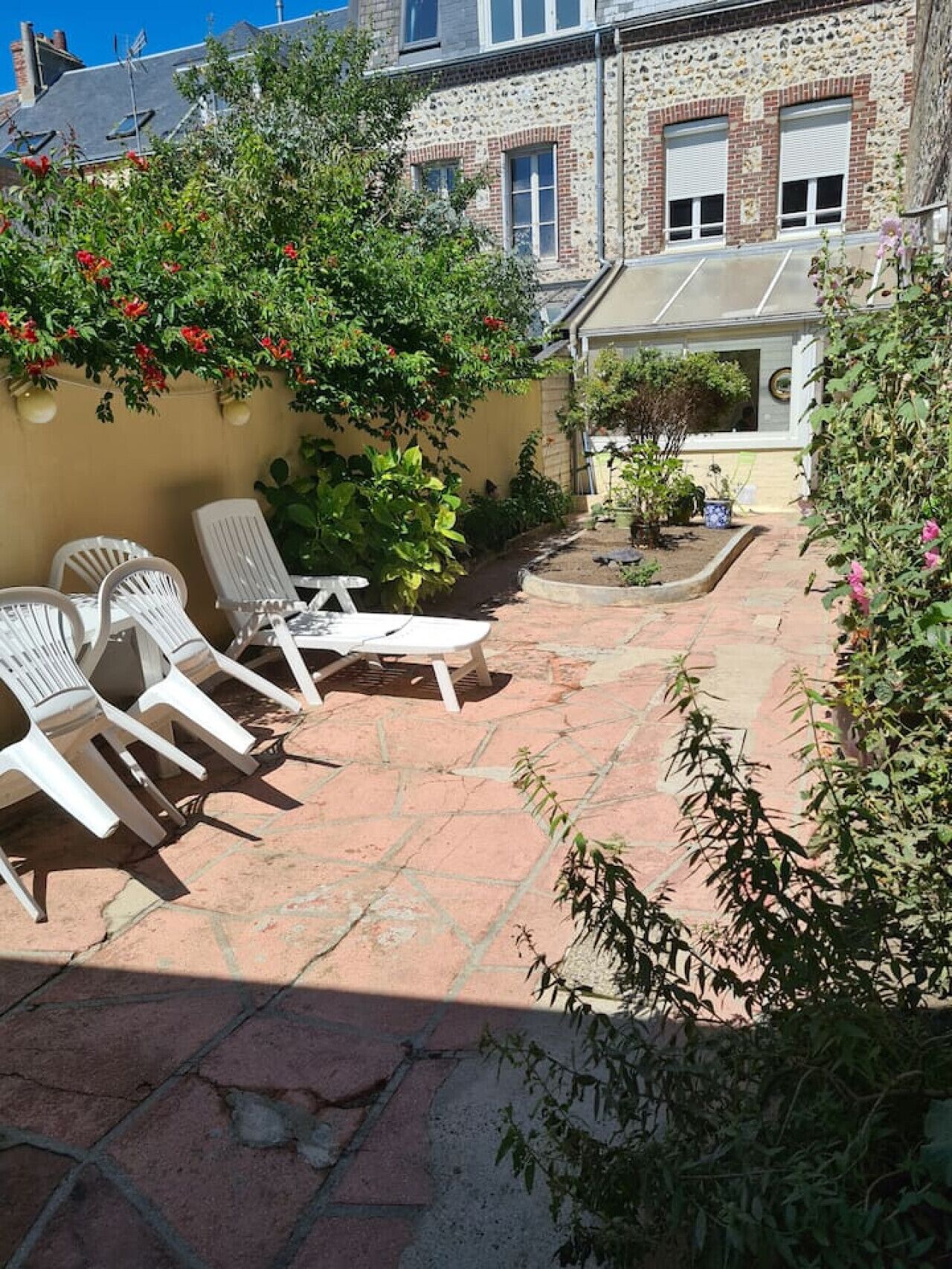 A patio with white lounge chairs, a small table, and a garden area with various plants and flowers.